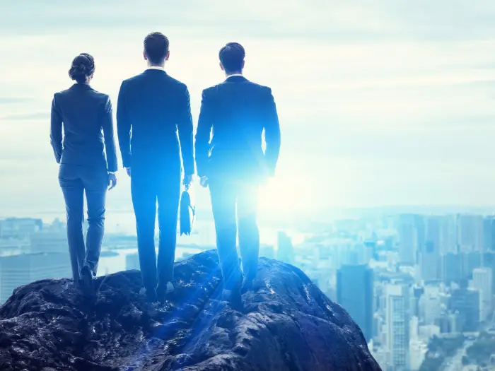 Three business professionals standing side-by-side on a mountain top looking over a large metropolitan area.