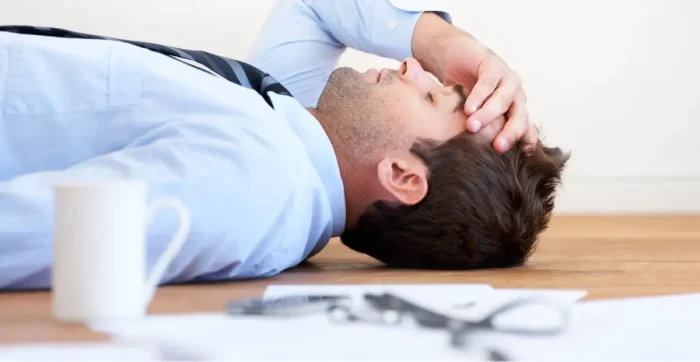 Stressed man in business attire lying on the floor with hand on forehead, surrounded by office documents and a coffee cup-illustrating burnout while studying for the CFA Level 1 exam.