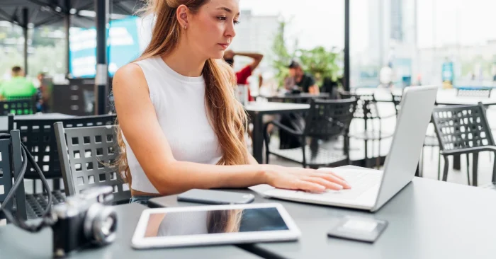 Working professional studying CFA materials on a laptop at an outdoor cafe, with camera and tablet beside her.