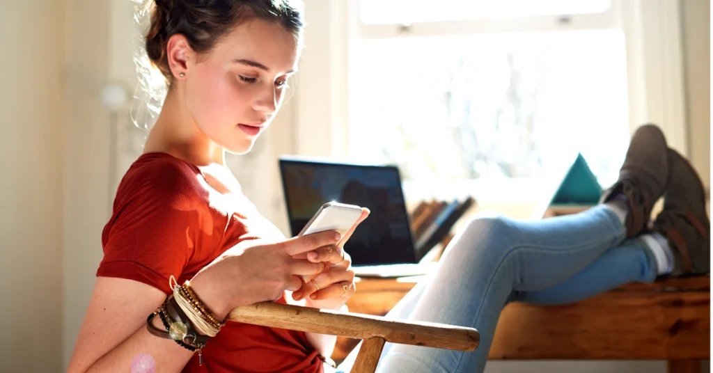 Young woman using her phone with a laptop nearby, illustrating a relaxed, personalized CFA weekly study plan setup.