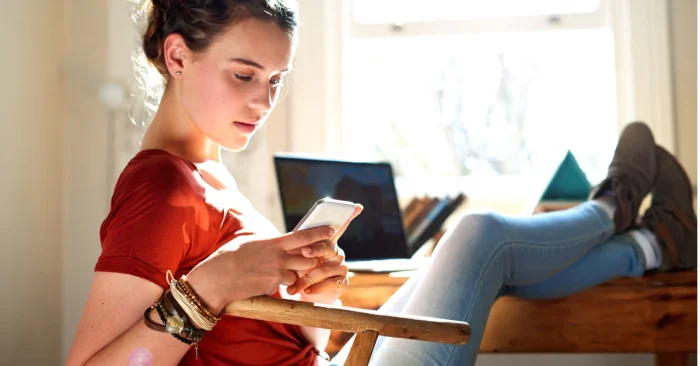 Young woman using her phone with a laptop nearby, illustrating a relaxed, personalized CFA weekly study plan setup.