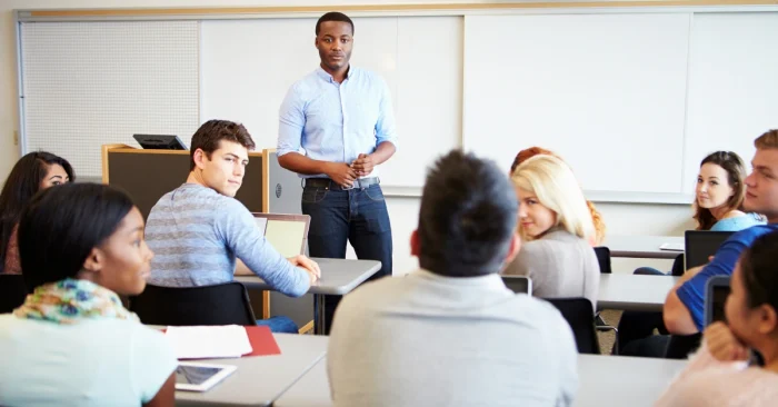 College students in a classroom setting, with one student standing and addressing the group representing full-time students discussing how to balance CFA exam prep with academic classes.